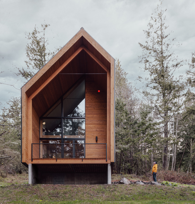  A modern A-frame house with large glass windows, featuring a warm wooden facade and a balcony, set against a backdrop of tall, slender trees in a forest setting, with a person in a yellow jacket standing outside, creating a contrast with the natural surroundings.