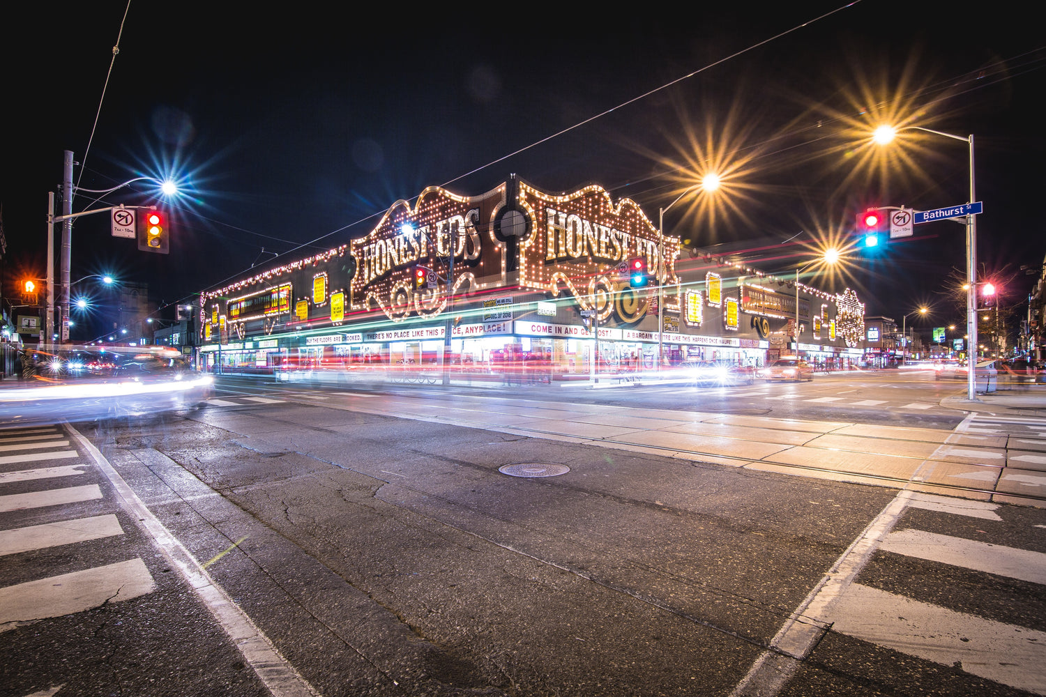  A bustling city intersection at night with the iconic, brightly lit Honest Ed's storefront. The long exposure captures the vibrant streaks of light from moving vehicles, against the static, illuminated signage, creating a lively urban atmosphere. Street signs and traffic lights add to the urban detail of the scene.