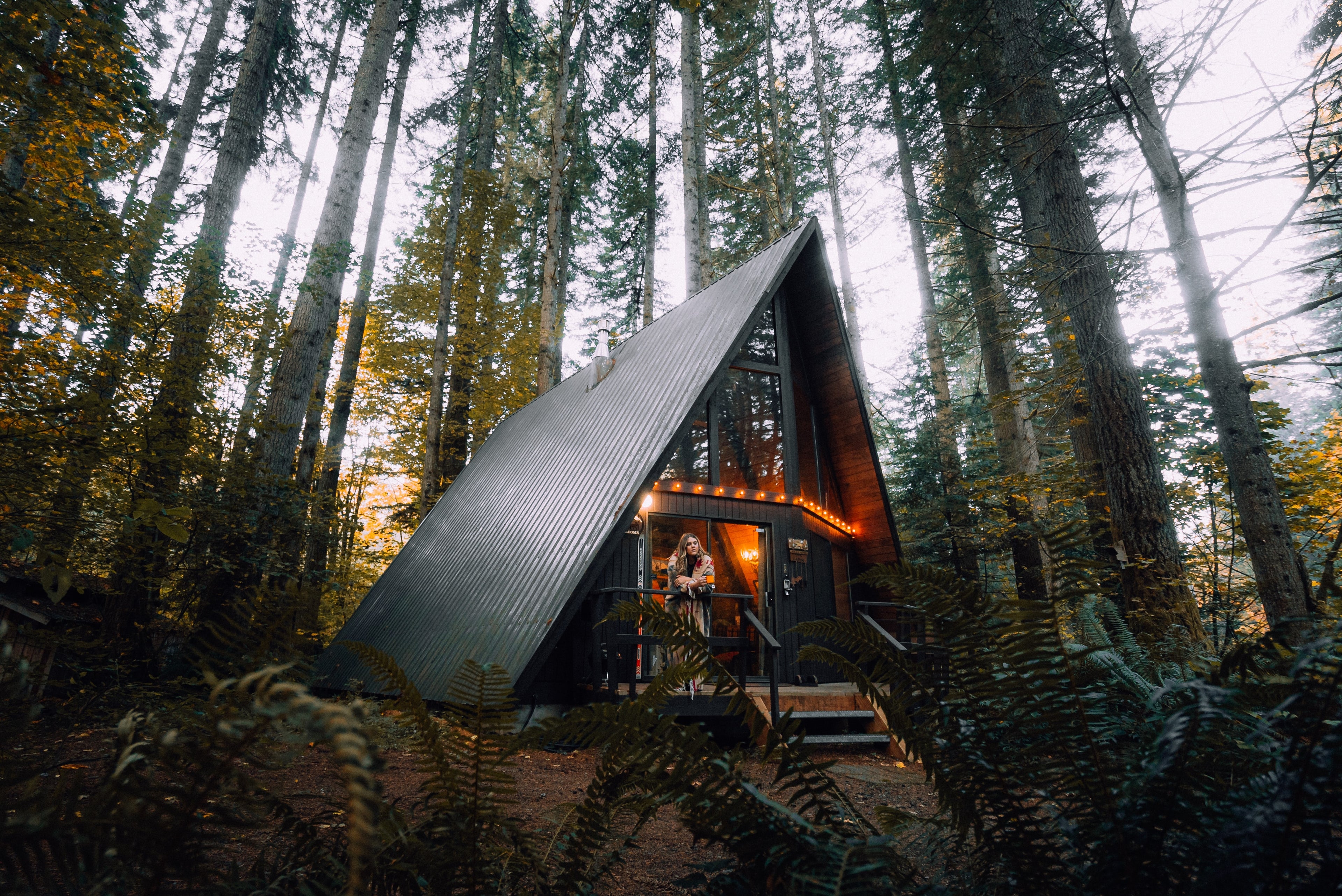  A cozy A-frame cabin nestled in a dense autumnal forest with warm lights illuminating the interior and a person standing on the front porch. Tall trees with yellowing leaves surround the cabin, evoking a serene, rustic retreat.