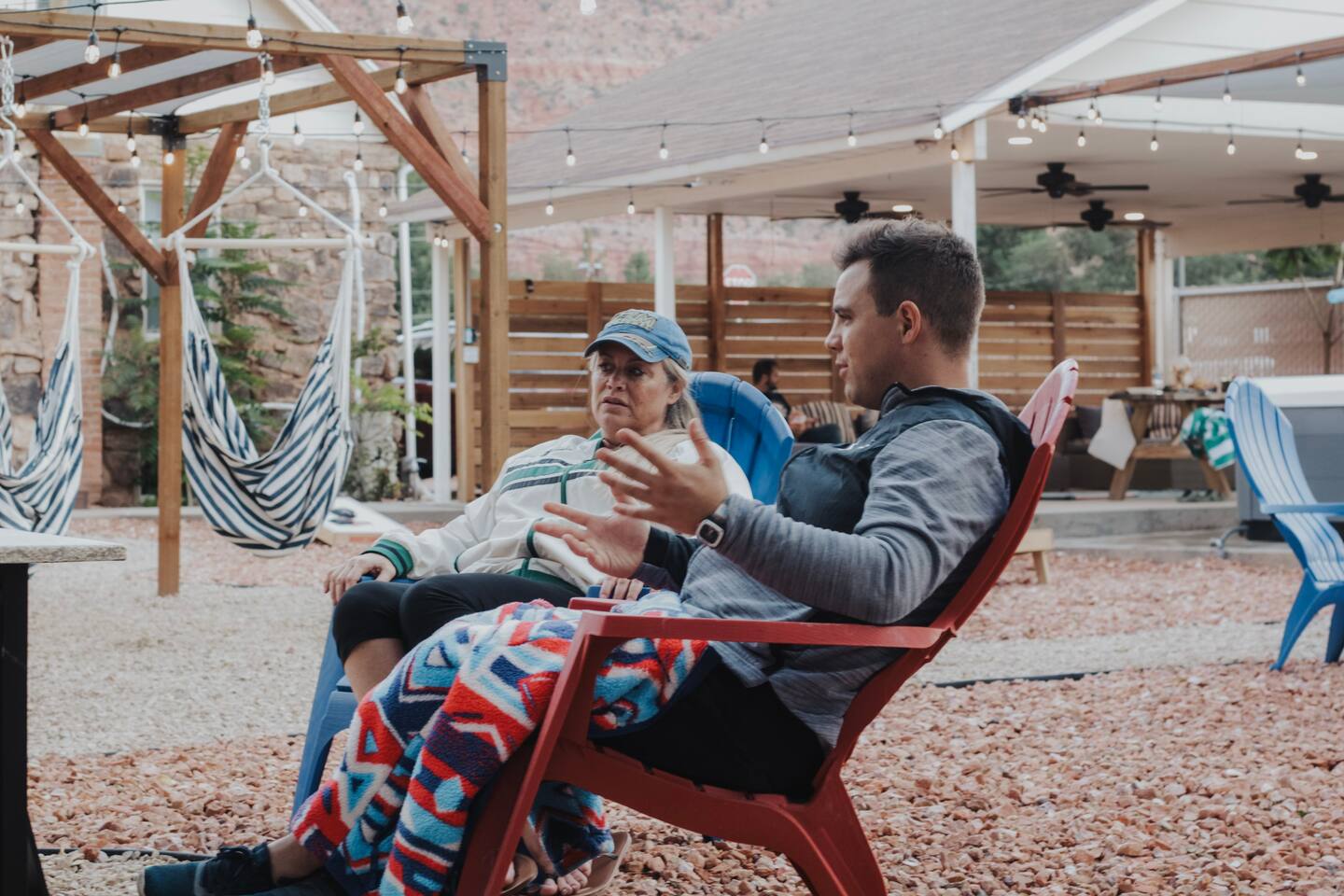  Two people engaged in a conversation while sitting on red Adirondack chairs in a patio area with a pebble ground cover, striped hammocks in the background, and a pergola with string lights above.