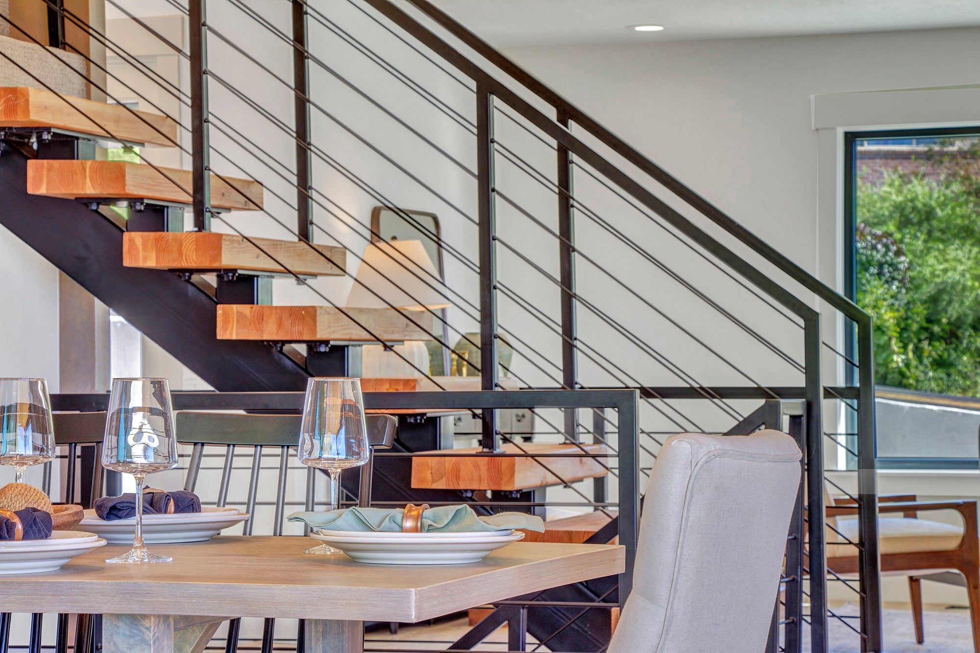 A modern dining area with a wooden table set with wine glasses and plates, adjacent to a sleek staircase with wooden steps and a black metal railing, all bathed in natural light from large windows.