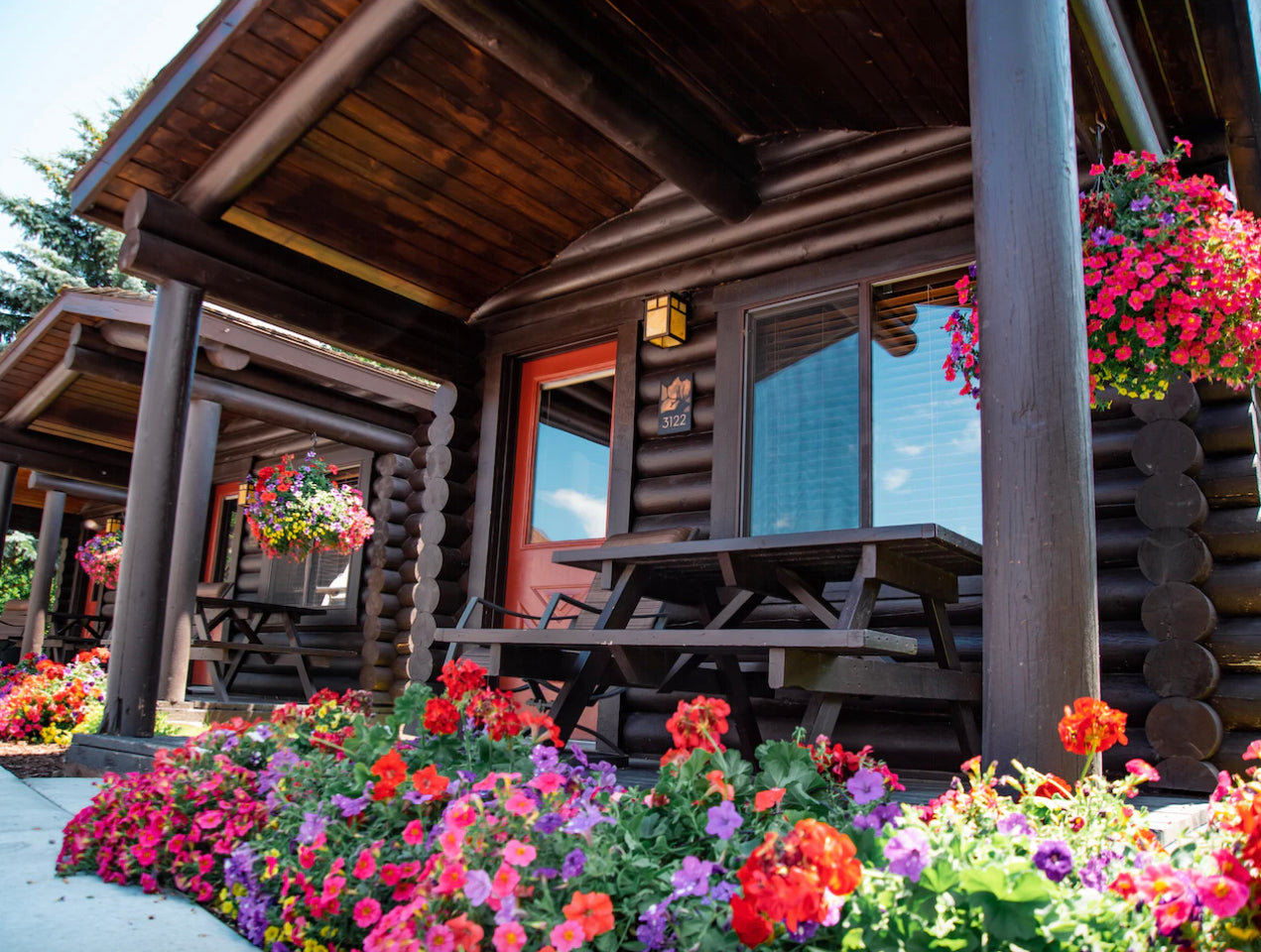  A log cabin exterior with vibrant flower boxes under the windows, a picnic table on the porch, and lush floral landscaping in the foreground, conveying a welcoming and rustic charm.