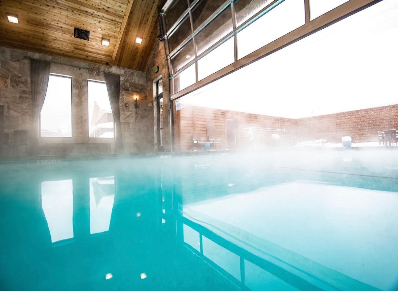  An indoor heated pool with steam rising off the water surface, featuring wood-paneled walls and ceiling, large windows allowing natural light, and a cozy, inviting atmosphere.