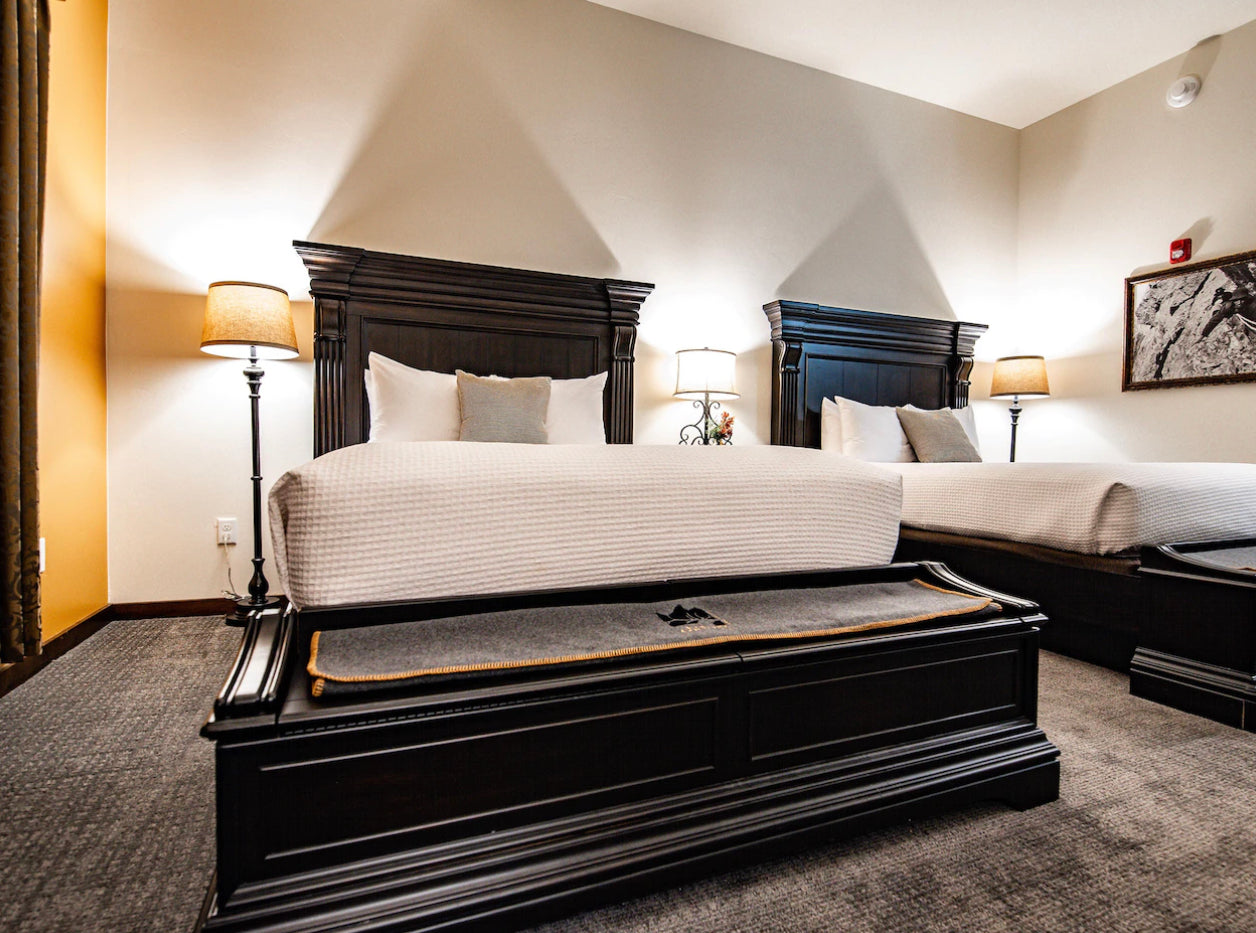  A hotel room featuring two large beds with dark wood headboards, crisp white bedding, and a warm, inviting ambiance accentuated by soft lighting from standing lamps and a neutral color scheme.