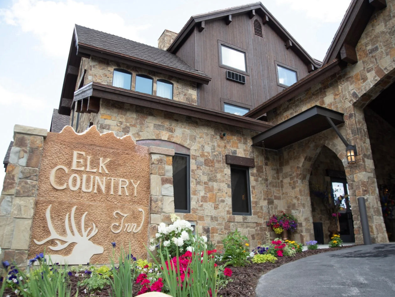  Exterior view of the 'Elk Country Inn' featuring a stone facade with a prominent sign, dark wood trims, a shingled roof, and beautifully landscaped garden beds filled with colorful flowers, under a partly cloudy sky.
