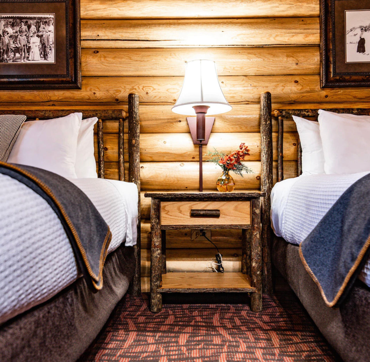  A rustic bedroom setting with two beds flanking a nightstand made of natural wood logs, a table lamp with a white shade, and historical framed pictures on a log cabin wall, evoking a cozy, traditional atmosphere.
