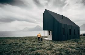  A minimalist dark-colored house stands in a vast, open landscape with a person in a yellow jacket next to it, suggesting solitude and tranquility. The overcast sky and distant mountains add to the remote and dramatic setting.