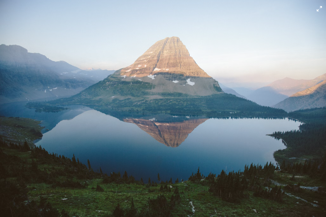  A serene landscape featuring a distinct, conical mountain reflected perfectly in the still waters of a lake below, with surrounding forested hills and a hazy sky hinting at early morning light.