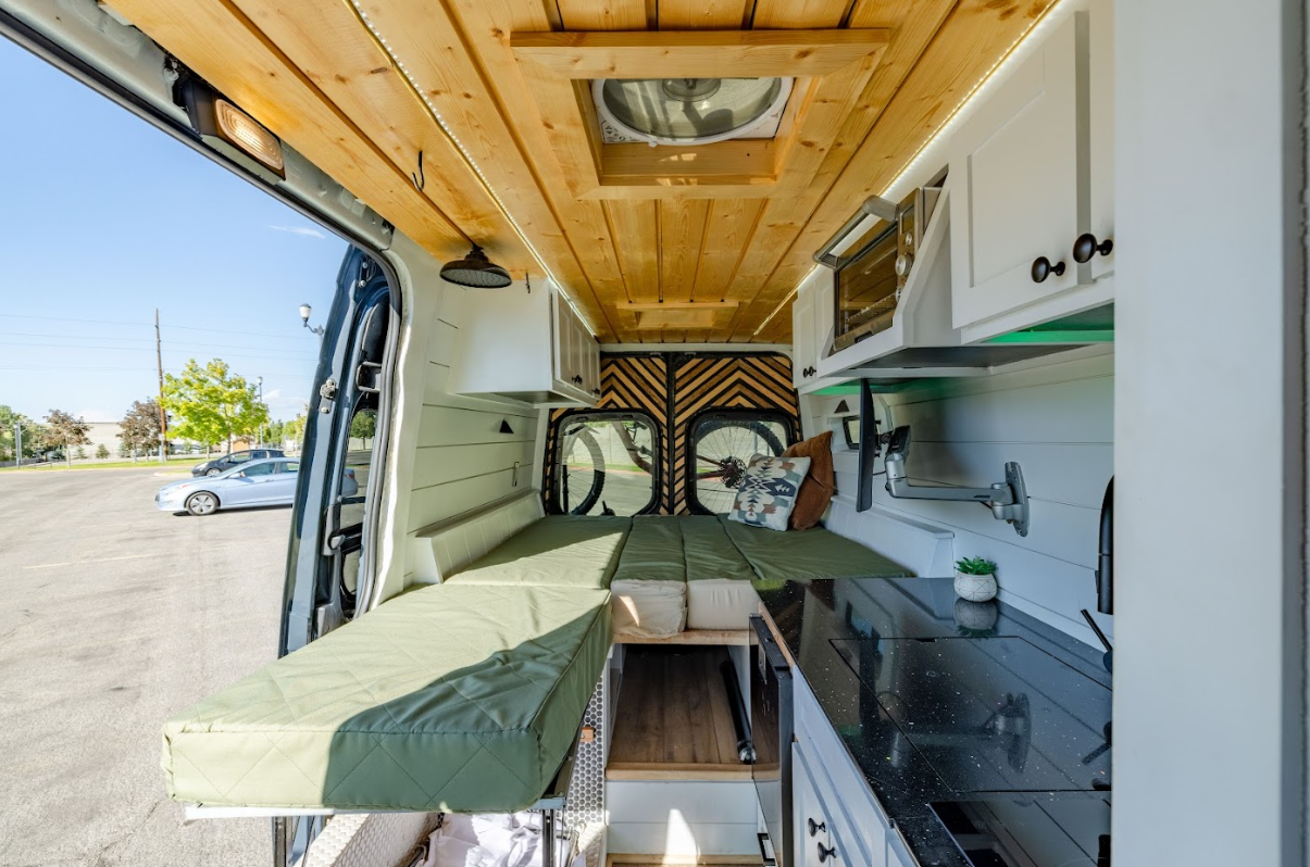  Interior of a modern camper van conversion featuring a cozy sleeping area, wooden ceiling, kitchenette with black countertops, and open rear doors, parked in a sunlit lot.