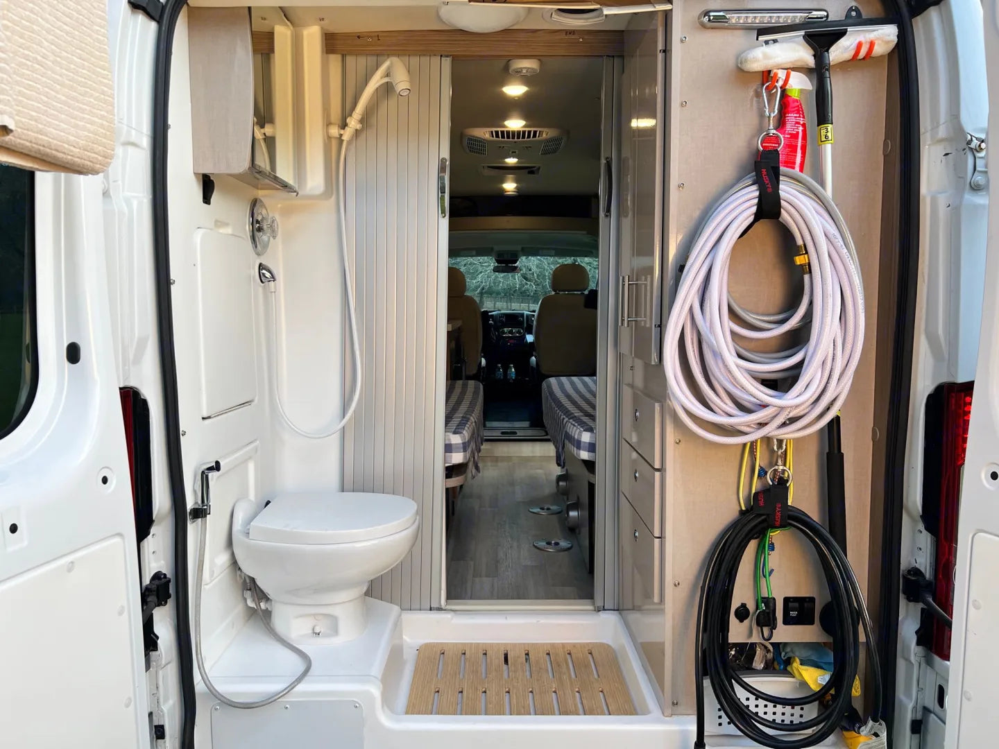  The back view of a camper van showing a compact and efficient layout with a white toilet, shower area, wooden flooring, and storage space including hoses and an extinguisher, with the interior living space visible in the distance.
