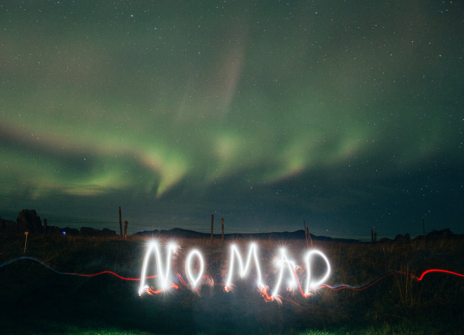  The photograph captures the ethereal beauty of the Northern Lights in a starlit sky, with an artistic light painting spelling out "NOMAD" in the foreground. The dynamic interplay of natural aurora borealis and human-made light art creates a vivid and memorable scene.