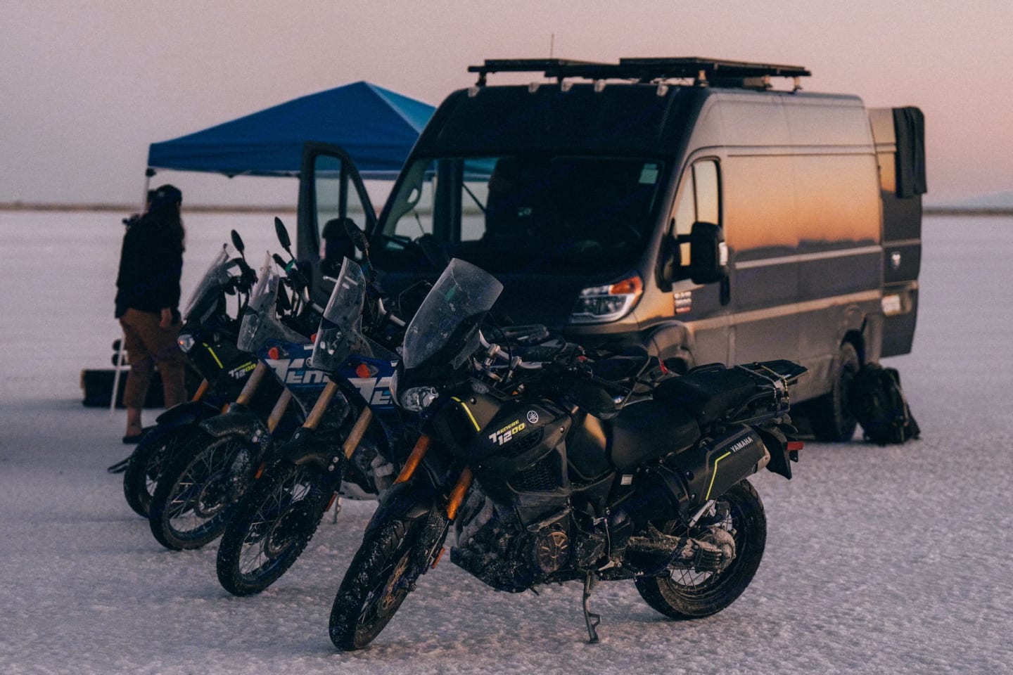 Adventure motorcycles parked on a flat, expansive salt flat at dusk, with a camper van and a blue canopy in the background, conveying a sense of exploration and travel.