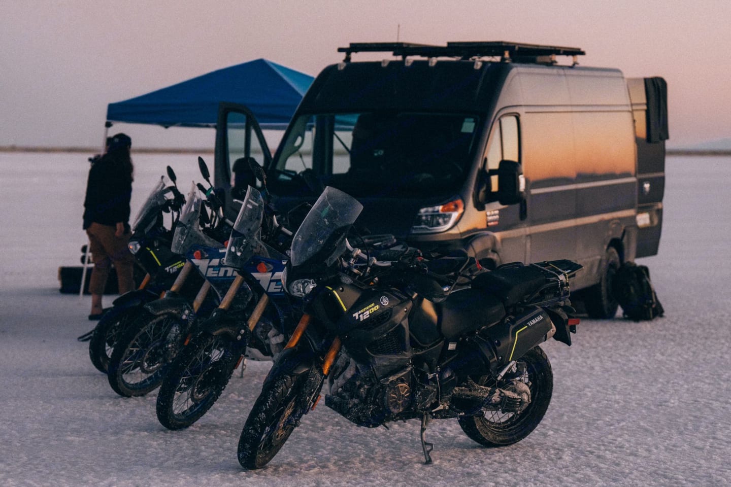 Adventure motorcycles parked next to a camper van on a snowy plain.