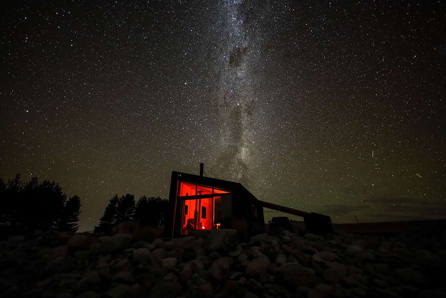  A solitary cabin illuminated with red light inside under a magnificent night sky filled with stars and the Milky Way, set against a backdrop of a dark landscape with silhouetted trees.